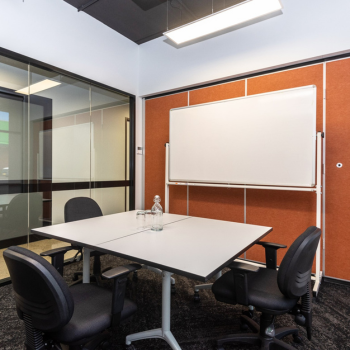 Modern meeting room with four chairs around a square table, glass walls, a whiteboard, and bottled water on the table.